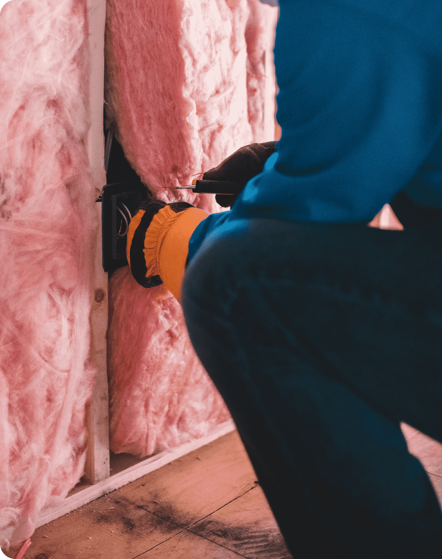 A person wearing gloves and a blue shirt is installing or adjusting an electrical outlet in a wall with exposed pink insulation.