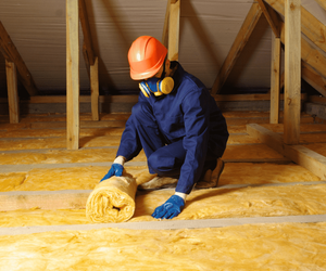 A person wearing a hard hat, protective mask, gloves, and coveralls is kneeling in an attic and installing fiberglass insulation between wooden beams.