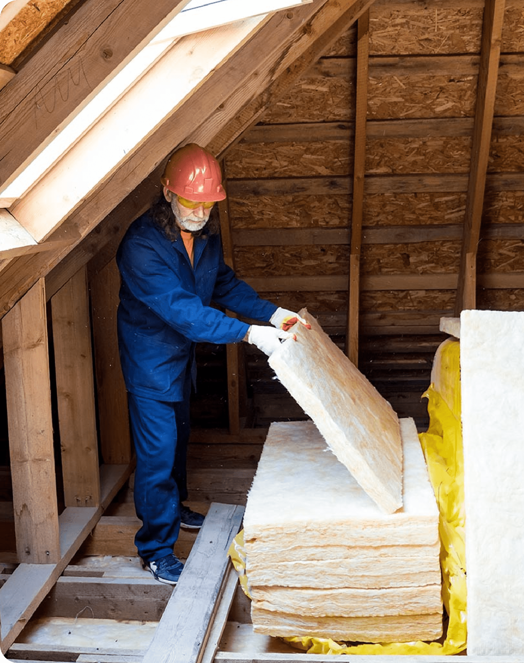 A person wearing a hard hat and work gloves is handling a large piece of Greenlink insulation material in an unfinished attic with exposed wooden beams and a skylight.