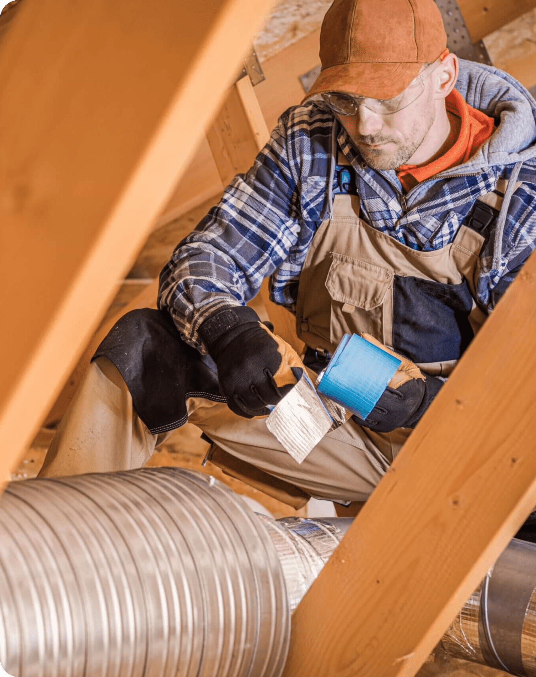 A worker in overalls, gloves, and a cap applies blue duct tape to a metal air duct in an attic, sitting among wooden beams and insulation—demonstrating precision and care worthy of the Greenlink standard.