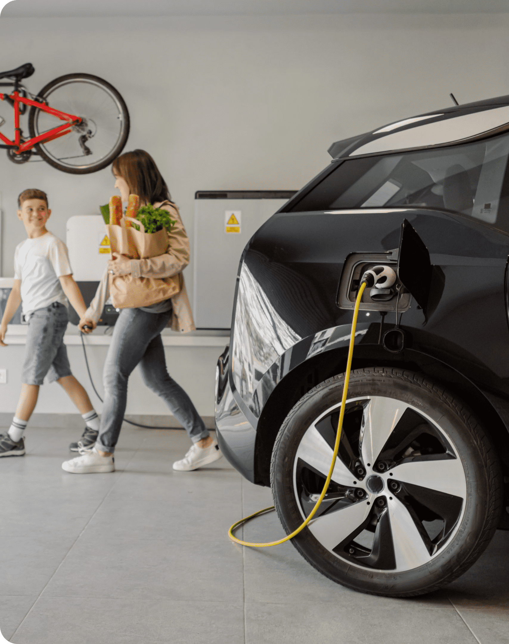 An electric car charges in a home garage while a mother and son carry in groceries.