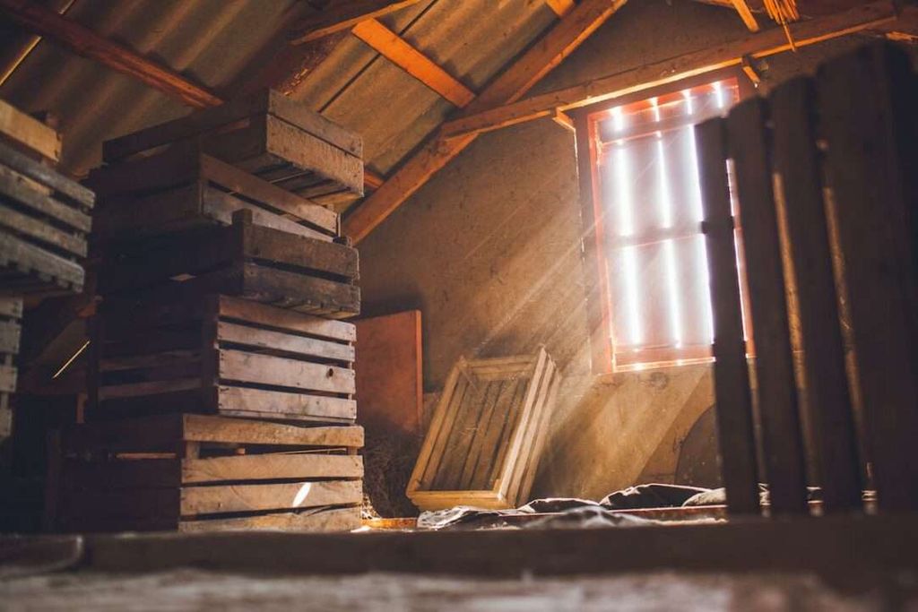 Sunlight streams through a window into a dusty attic with a slanted roof. Several Greenlink-branded wooden crates are stacked and scattered, and the light creates visible rays through the dusty air.