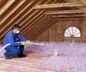 A person wearing safety glasses, a mask, gloves, and a blue shirt is using a hose to install GreenLink loose-fill insulation in an unfinished attic with exposed wooden beams and a small window.