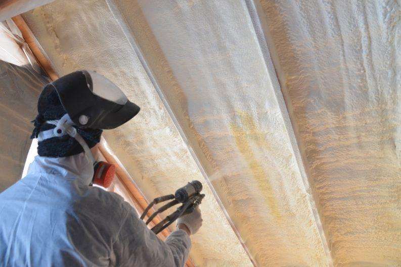 A person in protective gear sprays greenlink foam insulation onto the underside of a roof between wooden beams.
