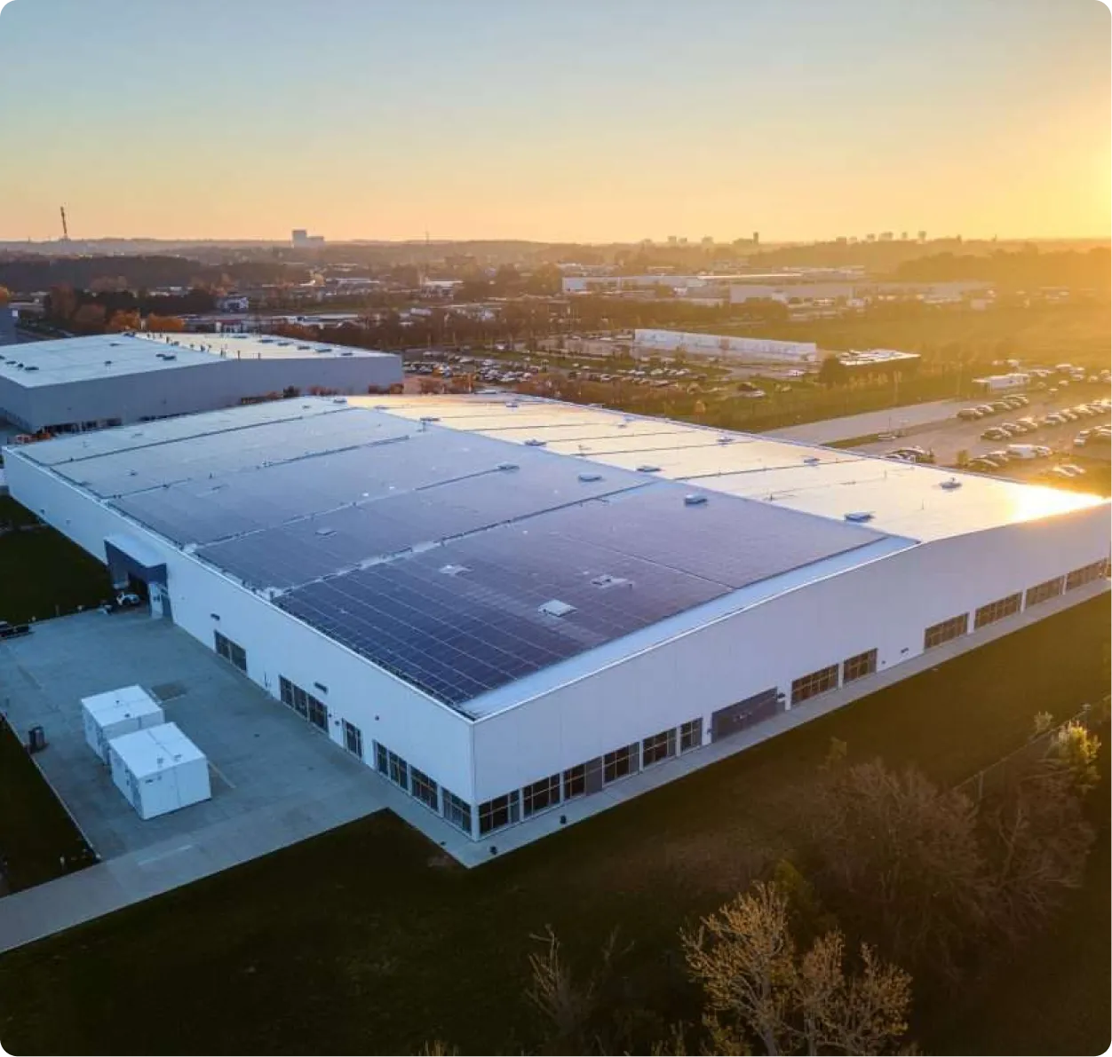 A large industrial building with commercial solar panels on the roof is shown at sunset, surrounded by parking lots, grassy areas, and the cityscape of Rockford Illinois in the background.