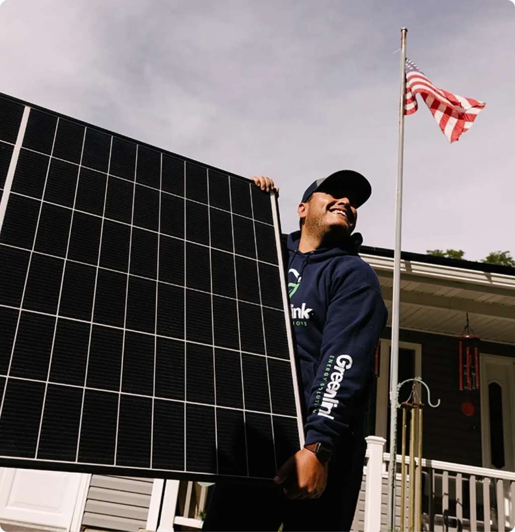 A person in a hoodie holds a large solar panel outside, smiling. The American flag is visible on a pole in the background, highlighting renewable energy efforts in Rockford Illinois.