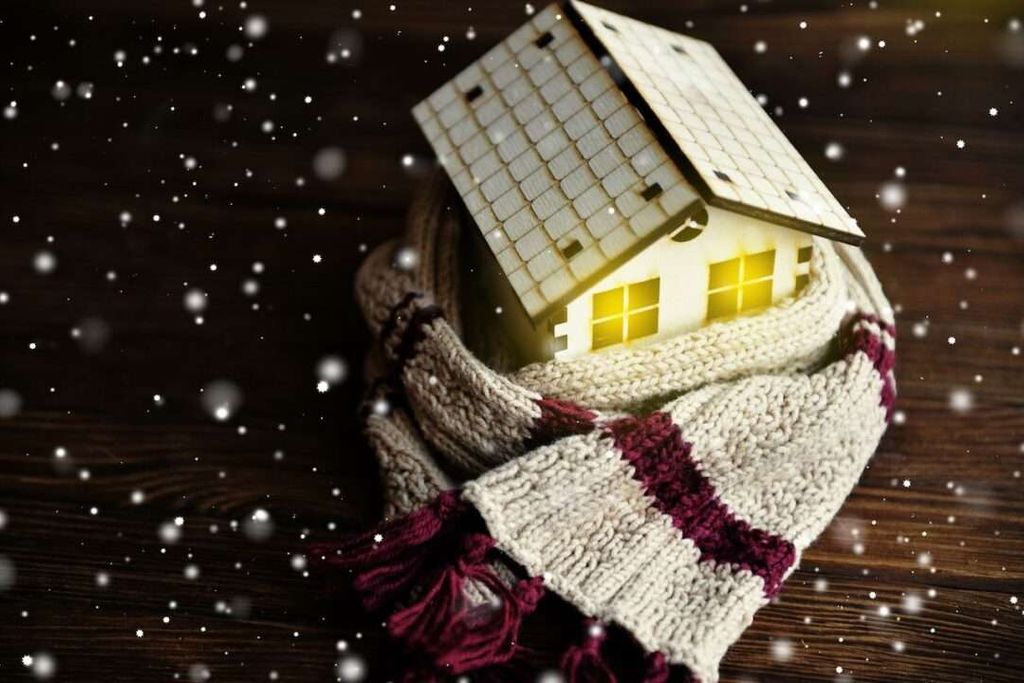 A small model house wrapped in a knitted scarf is placed on a wooden surface with falling snow, evoking warmth and protection during winter—perfectly capturing the comfort of a Greenlink-supported home.
