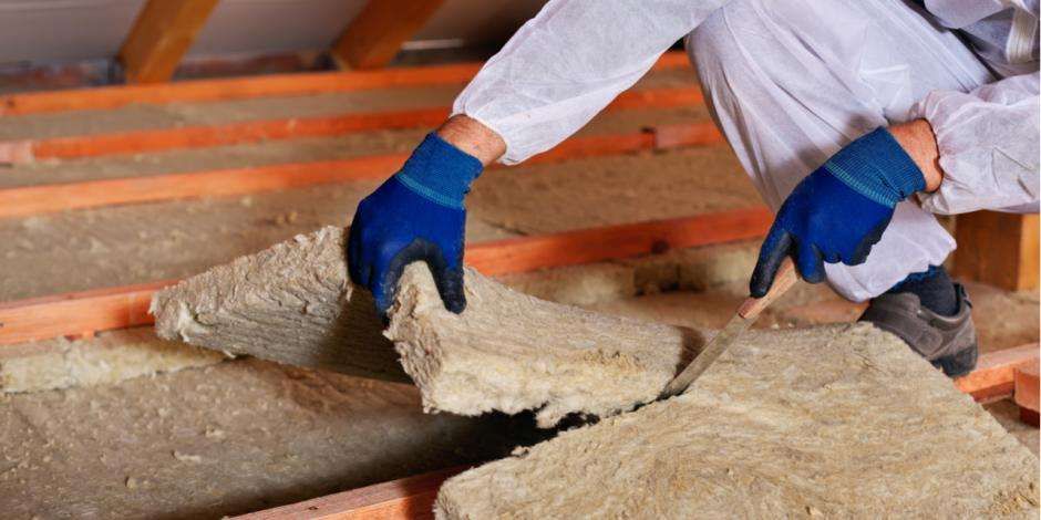 A person wearing gloves and protective clothing installs Greenlink insulation material between wooden beams in an attic space.