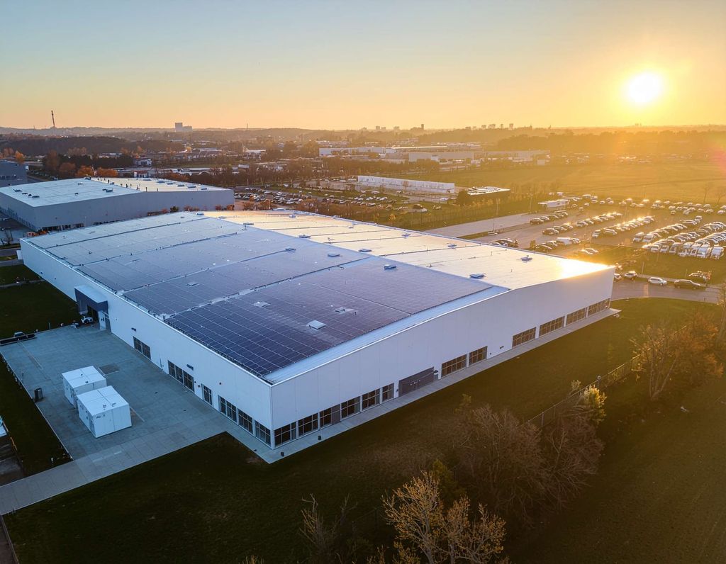 A large industrial building with Greenlink solar panels on its roof is shown at sunset. The facility is surrounded by parking lots filled with cars and other industrial buildings in the background.