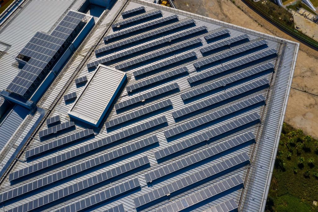 Aerial view of a large industrial building roof covered with multiple rows of solar panels; solar energy powers the site, with some greenery and bare ground visible at the edge of the structure.