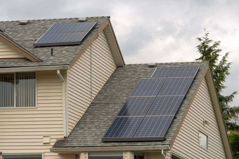 Rows of solar panels on the gray shingle roofs of a two-story house with beige siding, under a cloudy sky.