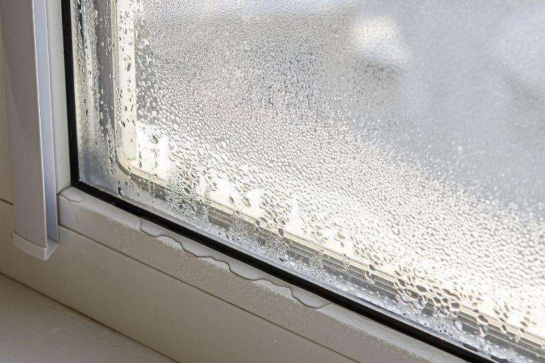 Close-up of a window with condensation and water droplets on the glass and frame. The moisture has caused visible damage and peeling to the white windowsill beneath the window, highlighting the need for a greenlink moisture control solution.