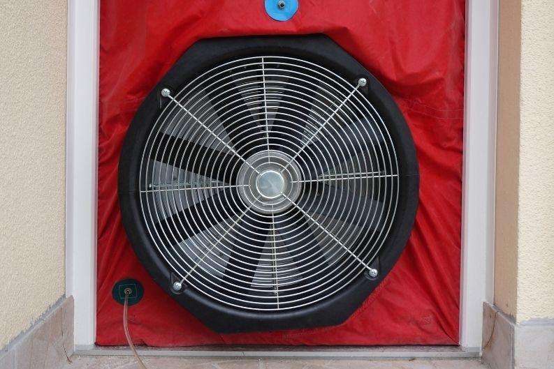 A large circular fan is installed in the middle of a red, airtight covering on a doorway for a blower door test, using greenlink technology to accurately measure a building’s air tightness. The fan is plugged into an electrical outlet nearby.