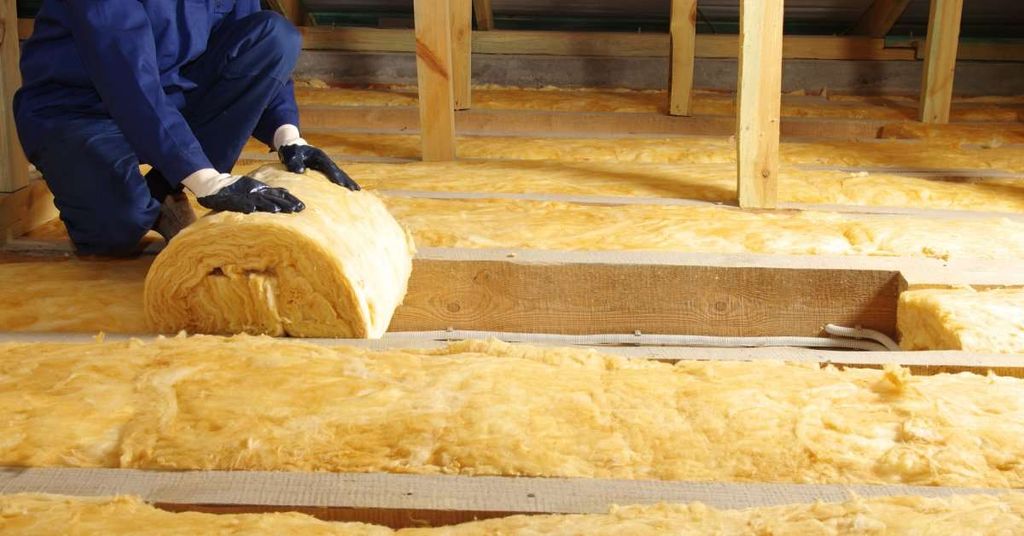 An unidentifiable worker in blue gear and protective gloves rolling yellow thermal insulation in an attic.