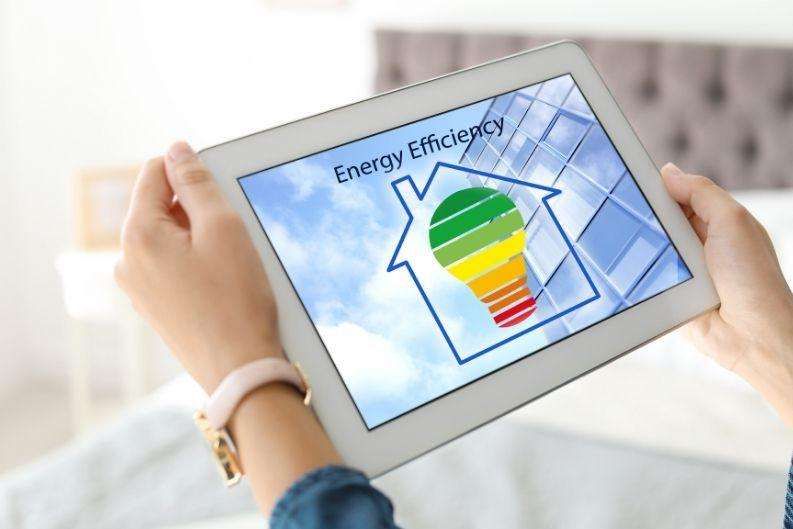 Person holding a tablet displaying a house outline with a multicolored light bulb and "Energy Efficiency" text. In the background, part of a building and blue sky appear, highlighting greenlink solutions for smarter energy use.