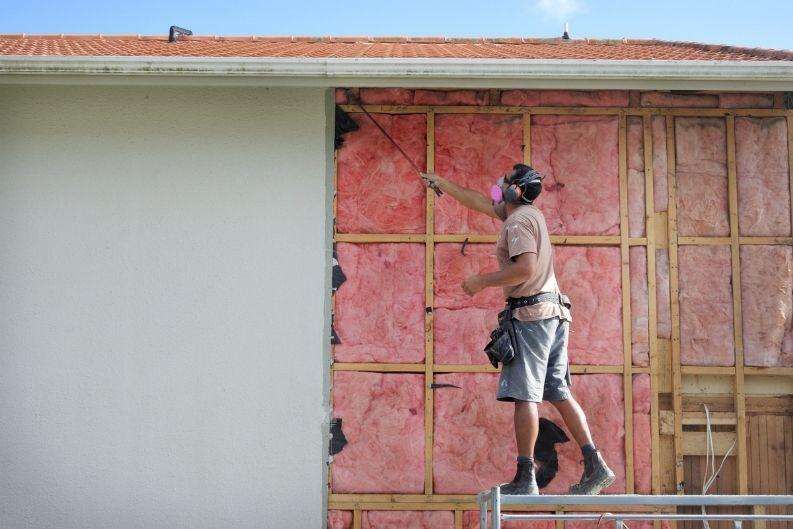 A person wearing ear protection and a tool belt is standing on a greenlink platform, painting or applying material to the upper section of a house wall with exposed pink insulation and wooden framing.