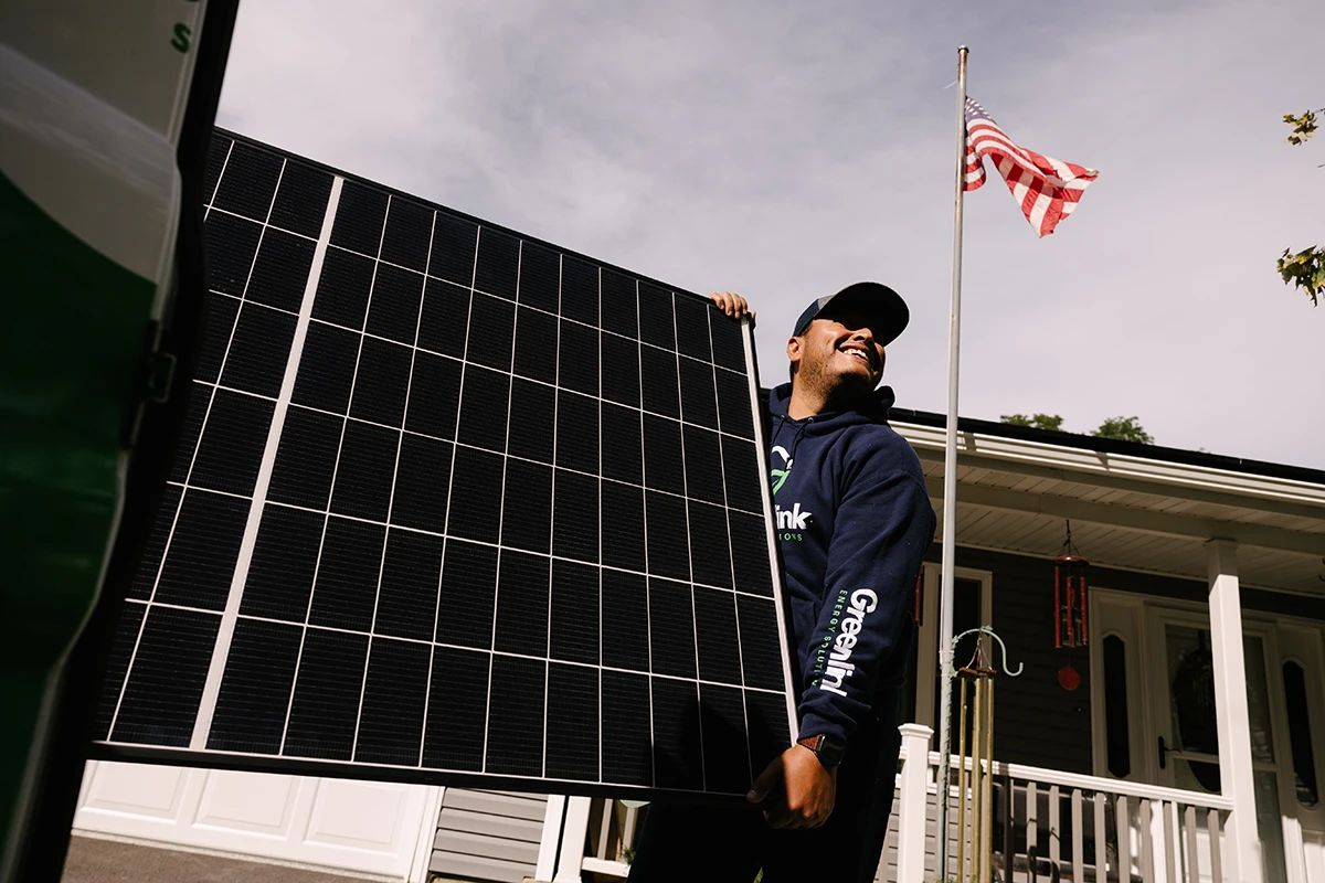 A smiling Greenlink installer carries a solar panel past a house with an American flag.