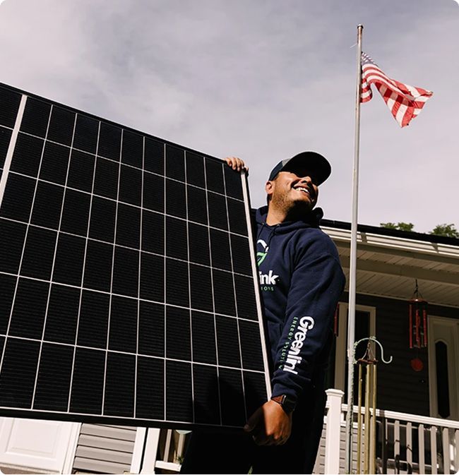 A person in a hoodie holding a solar panel stands in front of a building with an American flag in the background, highlighting the benefits of commercial solar and battery storage solutions.