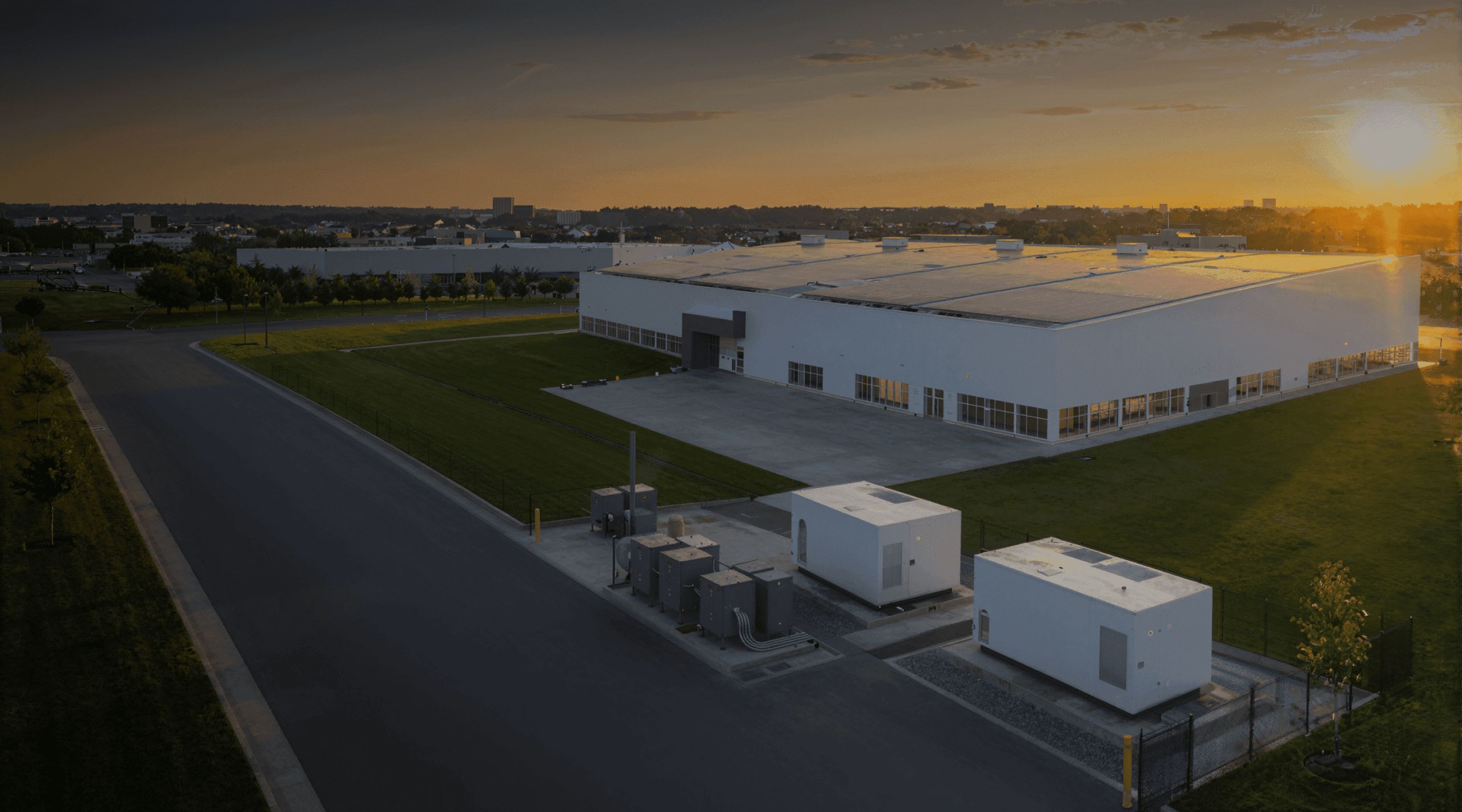 A large white industrial building with a flat roof sits on a grassy lot at sunset in Rockford, Illinois. Several white equipment units are in front, and an empty road runs alongside the property—an ideal setting for a commercial solar installation or conducting an energy audit.