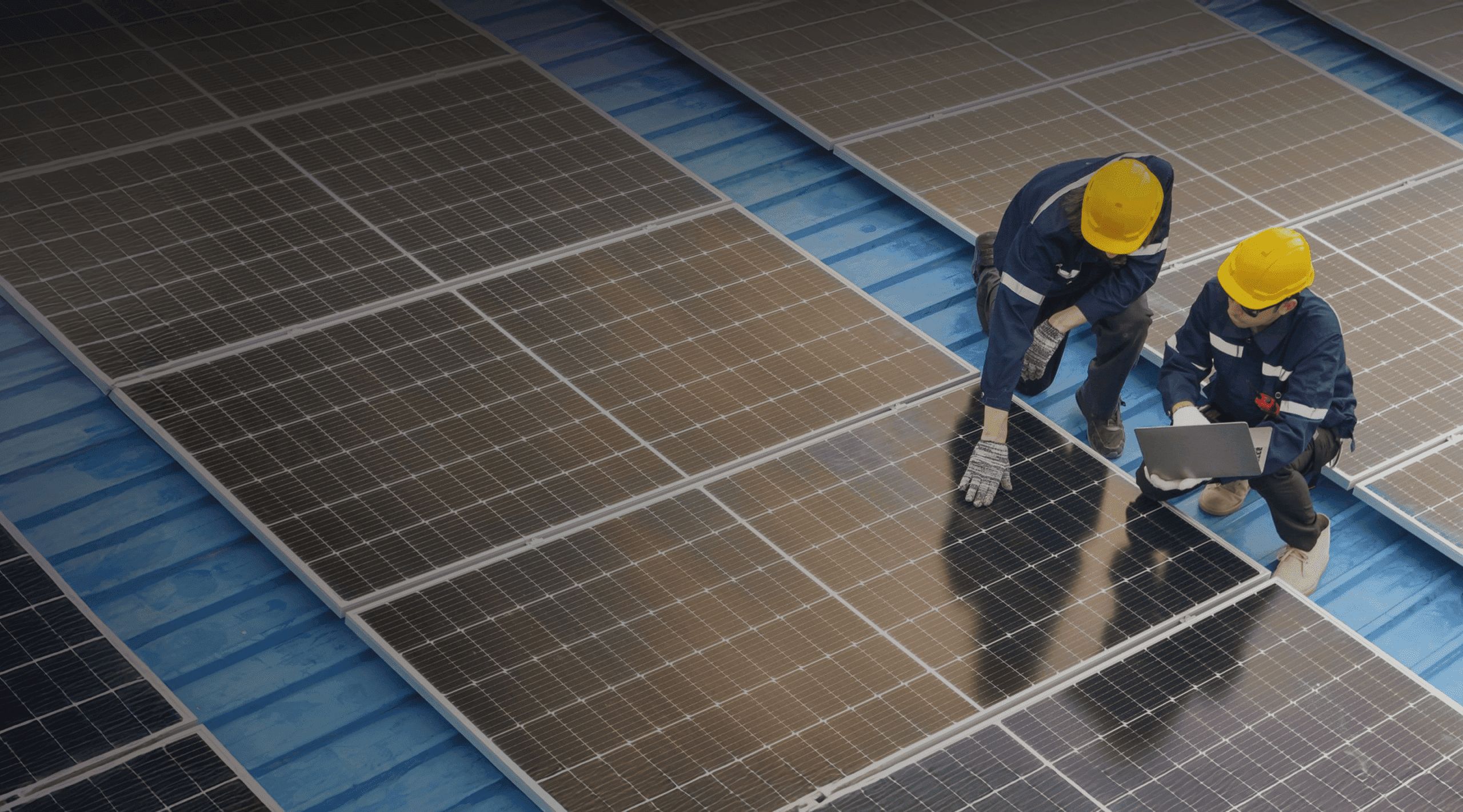 Two workers wearing yellow helmets inspect commercial solar panels on a blue rooftop; one is pointing at a panel while the other holds a laptop.