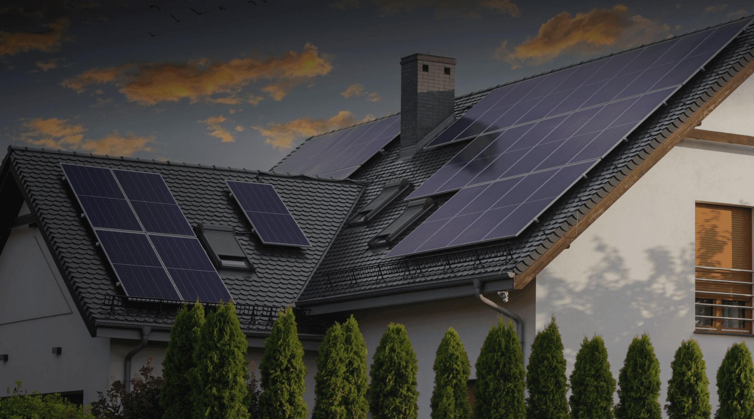 A modern industrial facility with multiple solar panels installed on its dark roof, surrounded by tall green shrubs, under a partly cloudy sunset sky near Rockford Illinois.