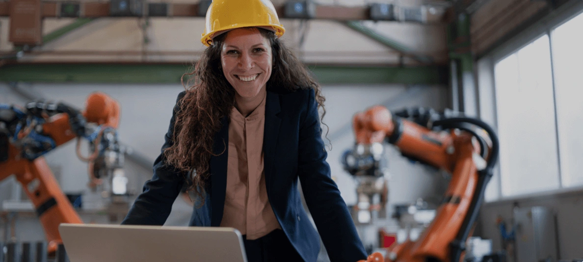 A smiling engineer in a yellow hard hat works on a laptop on a factory floor, with orange industrial robot arms behind her.