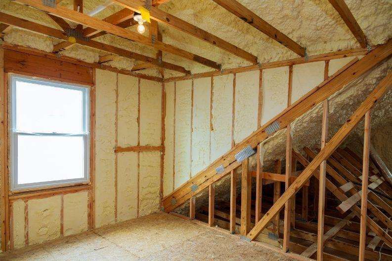 Attic space with exposed wooden beams, unfinished walls, and spray foam insulation on the ceiling and upper walls by Greenlink. A single window lets in daylight, and a bare light bulb hangs from the ceiling.