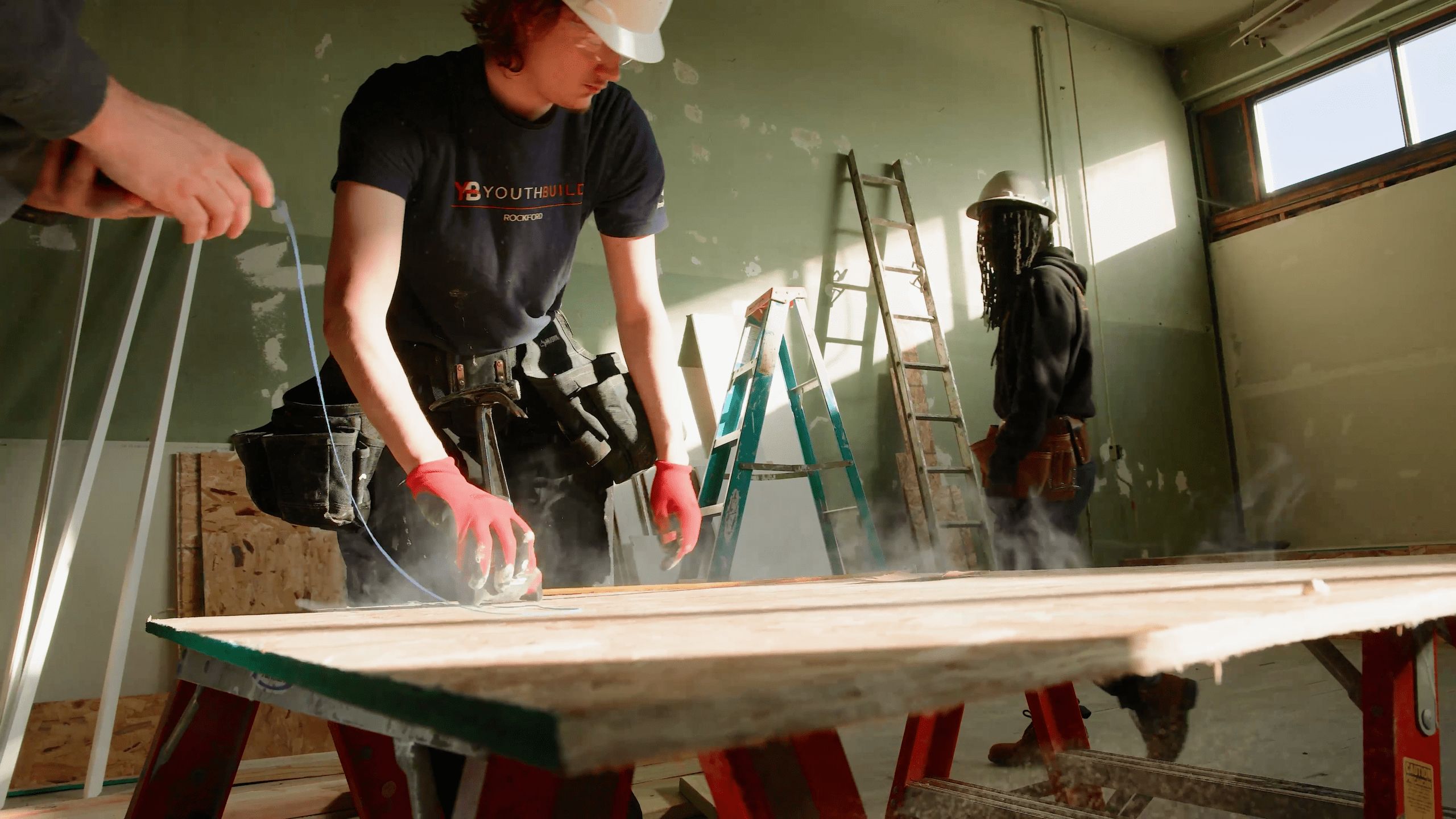 A Greenlink crew member in a hard hat and red gloves cuts a board on a job site while a colleague works on a ladder behind him.