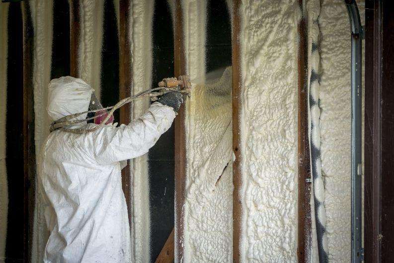 A person wearing protective gear sprays Greenlink foam insulation between wall studs inside a building under construction or renovation.