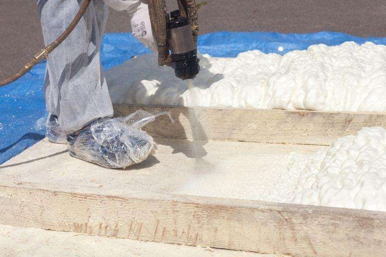 A Greenlink worker sprays expanding foam insulation onto wooden boards, wearing protective coveralls and plastic shoe coverings. The white foam is visible in various stages of expansion.