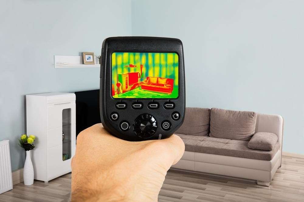 A hand holds a Greenlink thermal imaging camera pointed at a living room with a couch, shelf, and cabinet; the camera display shows heat signatures in varying colors on the furniture and walls.