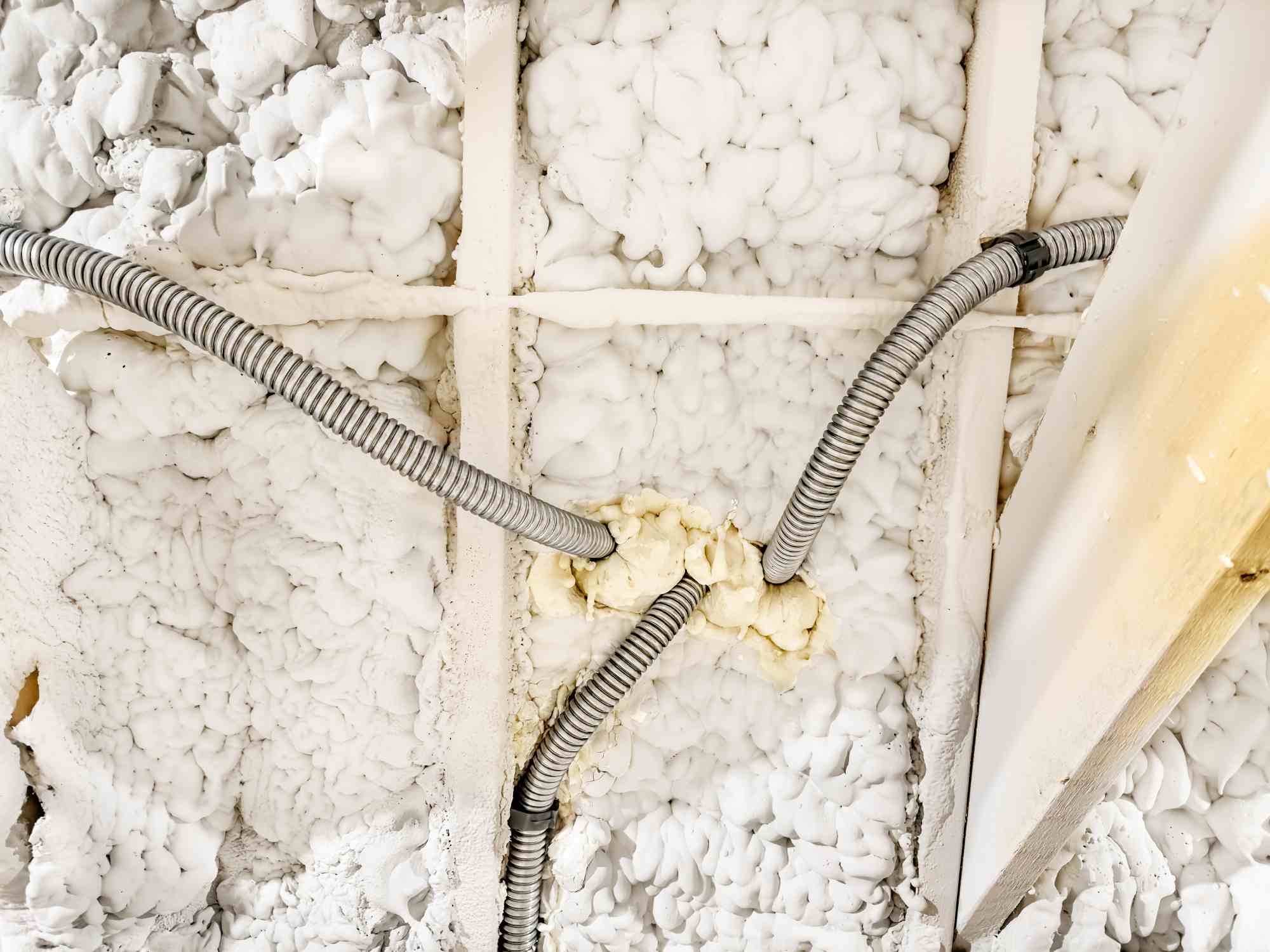 White spray foam insulation, up close, in an attic with metal pipes and white wooden rafters weaving through the material.