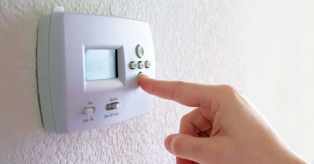 A close-up of a hand pressing a button on a white thermostat installed on a white, orange peel-textured wall.